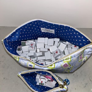 May include: Two zippered pouches, one large and one small, filled with white Mahjong tiles. The pouches have a blue polka dot lining and a light blue exterior with a floral and character design. The small pouch contains dice.