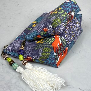 May include: Two navy blue zippered pouches with a forest and fox print. The pouches are stacked, with a white tassel and beaded zipper pull. The fabric features orange foxes, green foliage, and white accents.