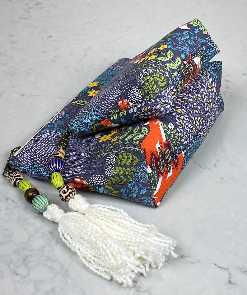 May include: Two navy blue zippered pouches with a forest and fox print. The pouches are stacked, with a white tassel and beaded zipper pull. The fabric features orange foxes, green foliage, and white accents.