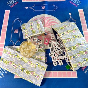 May include: A blue Mahjong game mat with matching accessories. The image shows a zippered pouch, a small coin purse, and a fabric wrap, all with a repeating pattern of Mahjong tiles. Dice, tiles, and a glass of ice are also present.