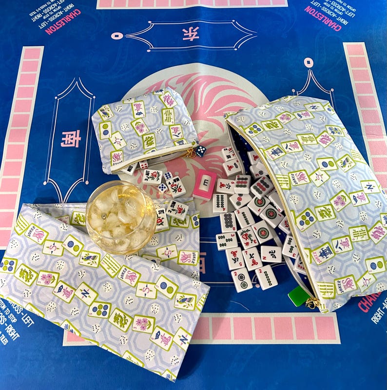 May include: A blue Mahjong game mat with matching accessories. The image shows a zippered pouch, a small coin purse, and a fabric wrap, all with a repeating pattern of Mahjong tiles. Dice, tiles, and a glass of ice are also present.