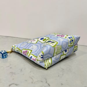 May include: A light blue fabric pouch with a gold zipper and a blue die attached. The pouch is patterned with playing card designs, flowers, and geometric shapes. The die is blue with white dots.