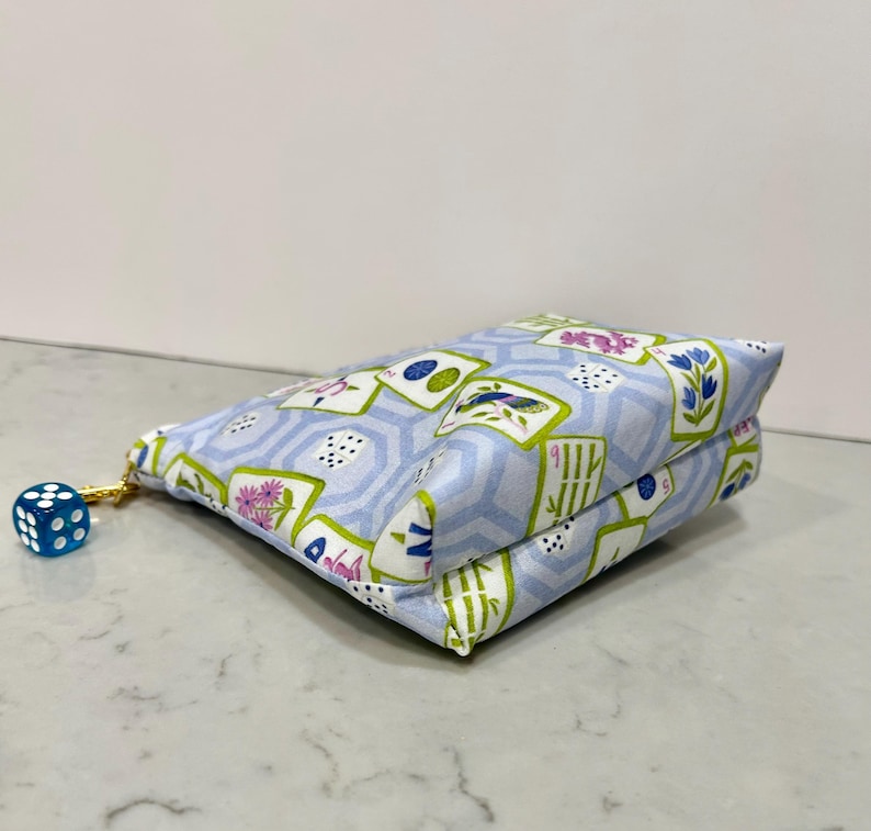 May include: A light blue fabric pouch with a gold zipper and a blue die attached. The pouch is patterned with playing card designs, flowers, and geometric shapes. The die is blue with white dots.