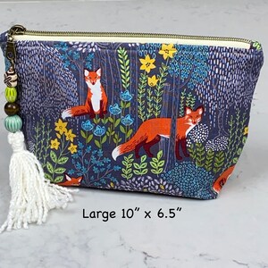 May include: A large, rectangular cosmetic bag with a forest and fox print. The bag is dark blue with a repeating pattern of orange foxes, yellow flowers, and blue foliage. It has a zipper closure and a tassel with beads. The bag measures 10 inches by 6.5 inches.