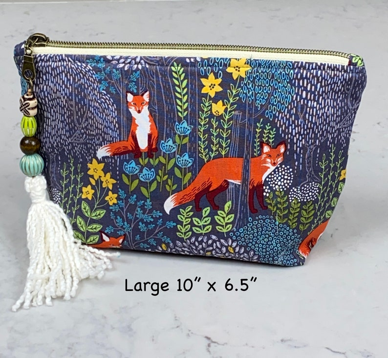May include: A large, rectangular cosmetic bag with a forest and fox print. The bag is dark blue with a repeating pattern of orange foxes, yellow flowers, and blue foliage. It has a zipper closure and a tassel with beads. The bag measures 10 inches by 6.5 inches.