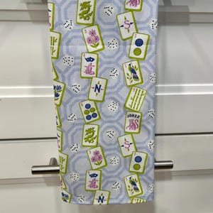 May include: A light blue and white kitchen towel with a repeating pattern of playing cards, dice, and geometric shapes. The cards feature various symbols and the word "JOKER". The towel is hanging on a silver bar.