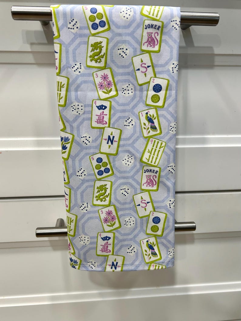 May include: A light blue and white kitchen towel with a repeating pattern of playing cards, dice, and geometric shapes. The cards feature various symbols and the word "JOKER". The towel is hanging on a silver bar.