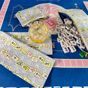 May include: A light blue Mahjong set with matching accessories, including a zippered pouch, a small coin purse, and a table runner. The set features a repeating pattern of Mahjong tiles, dice, and floral designs. A glass of iced tea sits in the center.