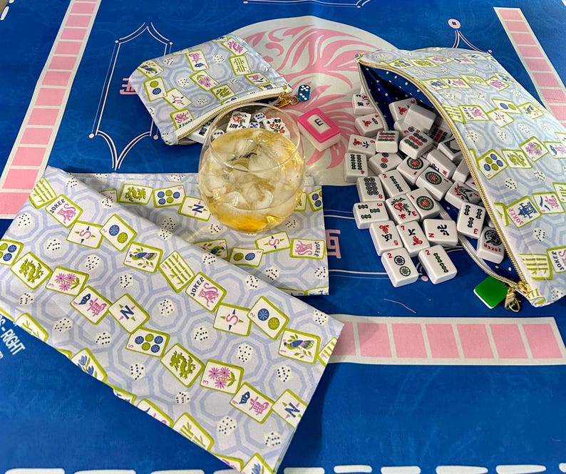 May include: A light blue Mahjong set with matching accessories, including a zippered pouch, a small coin purse, and a table runner. The set features a repeating pattern of Mahjong tiles, dice, and floral designs. A glass of iced tea sits in the center.