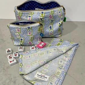 May include: Two light blue zippered cosmetic bags and a matching folded cloth featuring a Mahjong tile pattern in green, pink, and white. The bags have gold zippers and blue polka dot lining. Mahjong tiles and dice are scattered around.