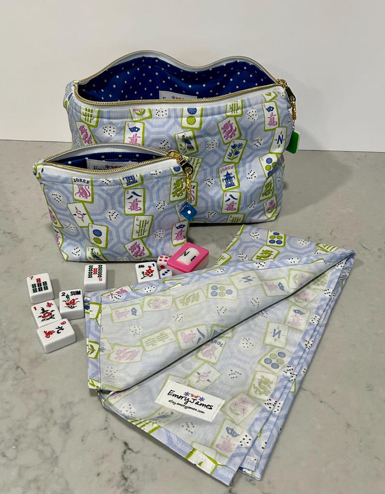 May include: Two light blue zippered cosmetic bags and a matching folded cloth featuring a Mahjong tile pattern in green, pink, and white. The bags have gold zippers and blue polka dot lining. Mahjong tiles and dice are scattered around.