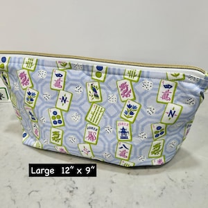 May include: A large, light blue cosmetic bag with a gold zipper and a Mahjong tile charm. The bag features a pattern of Mahjong tiles, dice, and floral designs. The bag measures 12 inches by 9 inches.