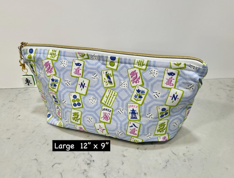 May include: A large, light blue cosmetic bag with a gold zipper and a Mahjong tile charm. The bag features a pattern of Mahjong tiles, dice, and floral designs. The bag measures 12 inches by 9 inches.