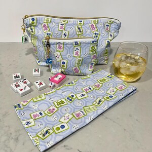 May include: A light blue cosmetic bag and matching small pouch with a gold zipper, featuring a Mahjong tile pattern. A matching napkin, Mahjong tiles, dice, and a glass of liquid with ice are also present.