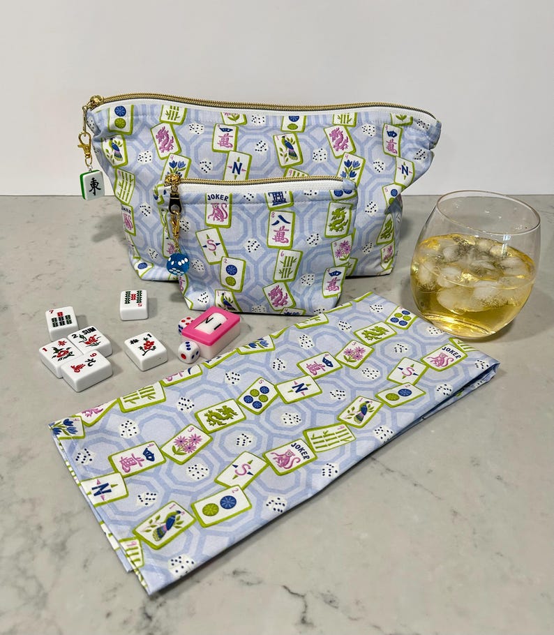May include: A light blue cosmetic bag and matching small pouch with a gold zipper, featuring a Mahjong tile pattern. A matching napkin, Mahjong tiles, dice, and a glass of liquid with ice are also present.