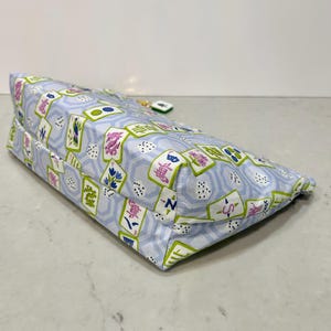 May include: A rectangular, light blue fabric bag with a repeating pattern of Mahjong tiles in green, pink, and blue. The bag has a zipper closure and a small charm attached to the zipper pull.