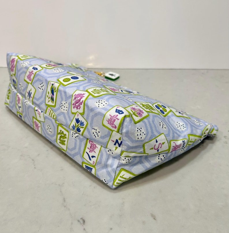 May include: A rectangular, light blue fabric bag with a repeating pattern of Mahjong tiles in green, pink, and blue. The bag has a zipper closure and a small charm attached to the zipper pull.