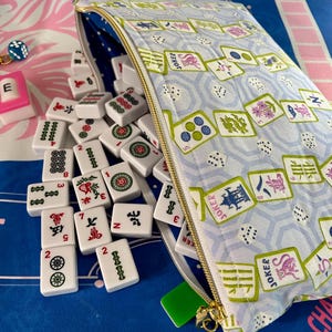 May include: A light blue Mahjong tile bag with a gold zipper, filled with white tiles featuring red, green, and black symbols. The bag is patterned with Mahjong tiles and dice. A few tiles are scattered outside the bag.