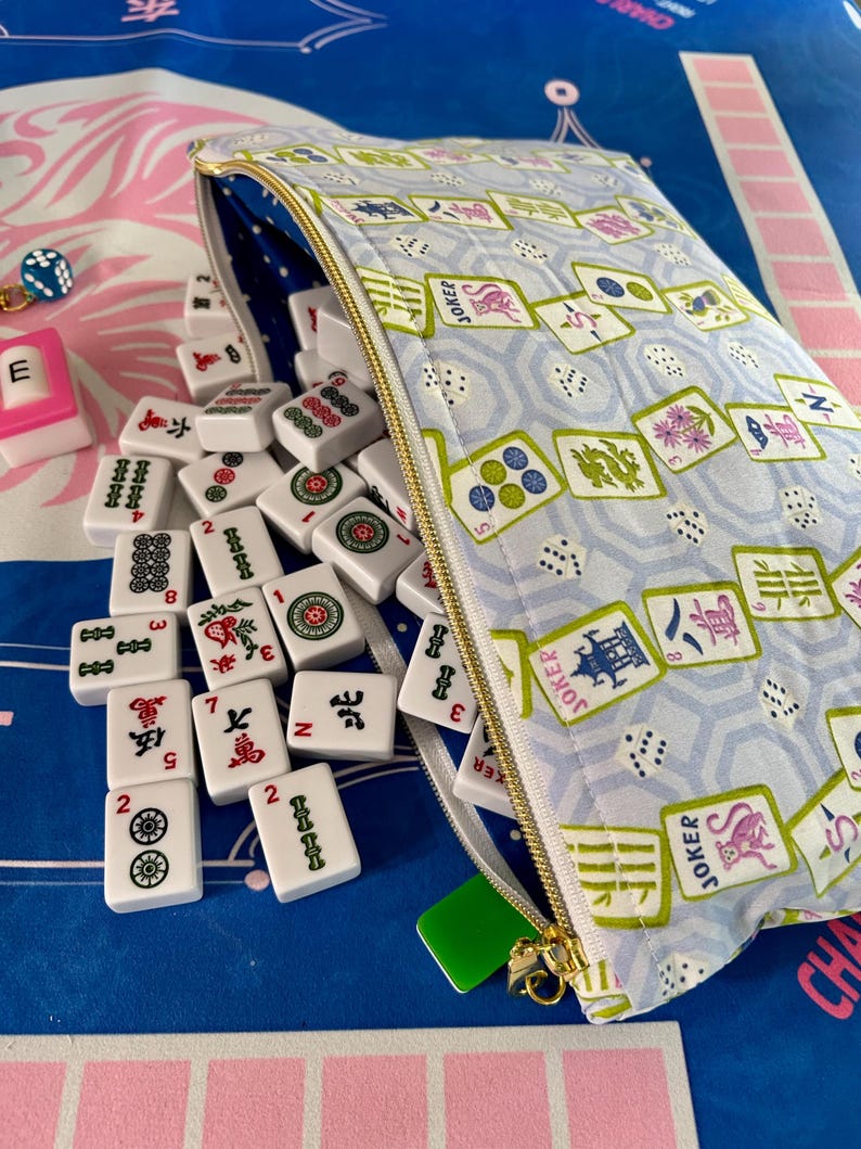 May include: A light blue Mahjong tile bag with a gold zipper, filled with white tiles featuring red, green, and black symbols. The bag is patterned with Mahjong tiles and dice. A few tiles are scattered outside the bag.