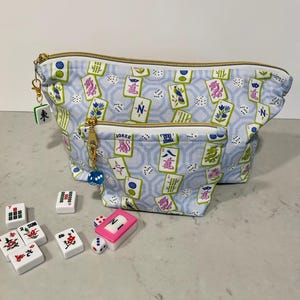May include: A light blue Mahjong-themed cosmetic bag with a gold zipper and a matching charm. The bag features a pattern of Mahjong tiles and dice. Several Mahjong tiles and dice are scattered in front of the bag.