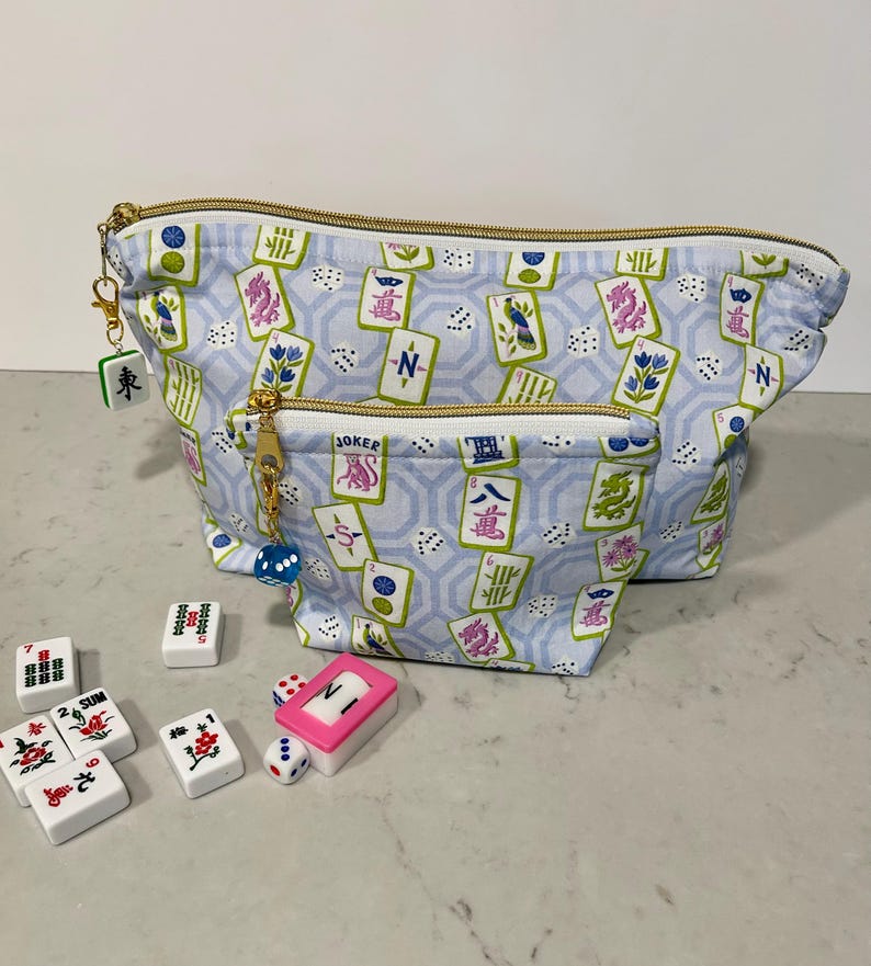 May include: A light blue Mahjong-themed cosmetic bag with a gold zipper and a matching charm. The bag features a pattern of Mahjong tiles and dice. Several Mahjong tiles and dice are scattered in front of the bag.
