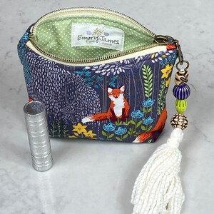 May include: A small, open zippered pouch with a forest scene featuring a fox, blue and yellow flowers, and trees. The pouch has a white tassel and beaded charm. A silver tube is next to the pouch. The interior lining is green with white polka dots.