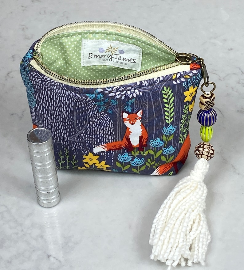 May include: A small, open zippered pouch with a forest scene featuring a fox, blue and yellow flowers, and trees. The pouch has a white tassel and beaded charm. A silver tube is next to the pouch. The interior lining is green with white polka dots.