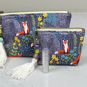 May include: Two zippered pouches with a forest fox print. The pouches feature a dark blue background with a repeating pattern of trees, flowers, and foxes. Each pouch has a gold zipper and a beaded tassel. A silver lipstick tube is in front of the pouches.