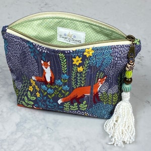 May include: A zippered pouch with a forest scene featuring foxes, flowers, and foliage in shades of blue, orange, and green. The interior is light green with white polka dots. A white tassel and beaded zipper pull add detail. The label reads "Emory James Fine Linens."