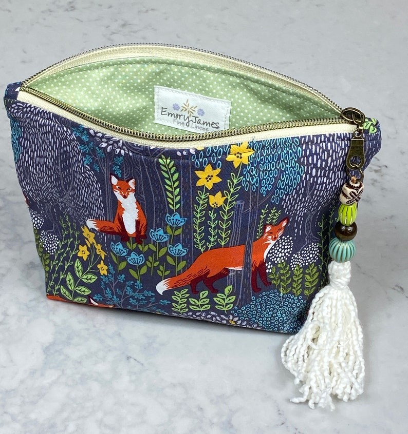 May include: A zippered pouch with a forest scene featuring foxes, flowers, and foliage in shades of blue, orange, and green. The interior is light green with white polka dots. A white tassel and beaded zipper pull add detail. The label reads "Emory James Fine Linens."