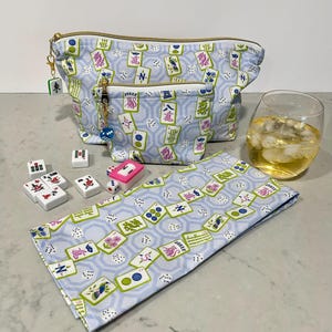 May include: A light blue cosmetic bag and matching small pouch with a gold zipper, featuring a Mahjong tile pattern. A matching napkin, Mahjong tiles, dice, and a glass of liquid with ice are also present.