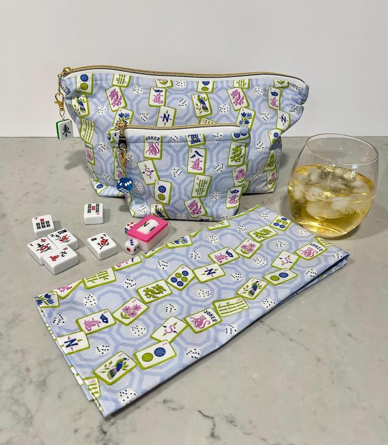 May include: A light blue cosmetic bag and matching small pouch with a gold zipper, featuring a Mahjong tile pattern. A matching napkin, Mahjong tiles, dice, and a glass of liquid with ice are also present.