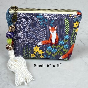 May include: A small, zippered pouch with a forest scene featuring a fox, flowers, and trees. The pouch is approximately 6 inches by 5 inches. It has a white tassel and beaded zipper pull. The background is navy blue.
