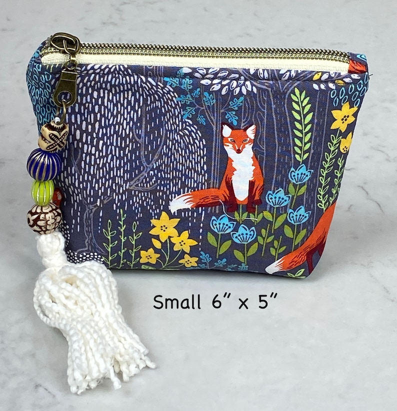 May include: A small, zippered pouch with a forest scene featuring a fox, flowers, and trees. The pouch is approximately 6 inches by 5 inches. It has a white tassel and beaded zipper pull. The background is navy blue.