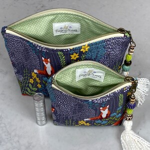 May include: Two open zippered cosmetic bags with a forest fox print. The bags have a navy blue background with a forest scene featuring foxes, trees, and flowers. The interior is light green with white polka dots. One bag has a white tassel and beaded zipper pull.