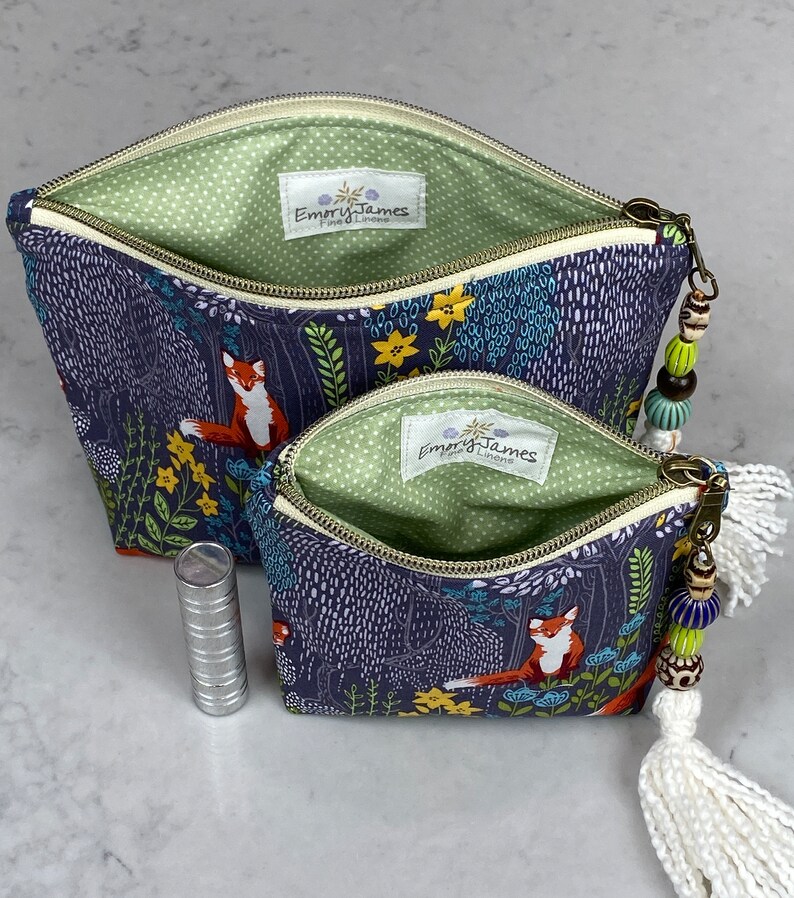 May include: Two open zippered cosmetic bags with a forest fox print. The bags have a navy blue background with a forest scene featuring foxes, trees, and flowers. The interior is light green with white polka dots. One bag has a white tassel and beaded zipper pull.