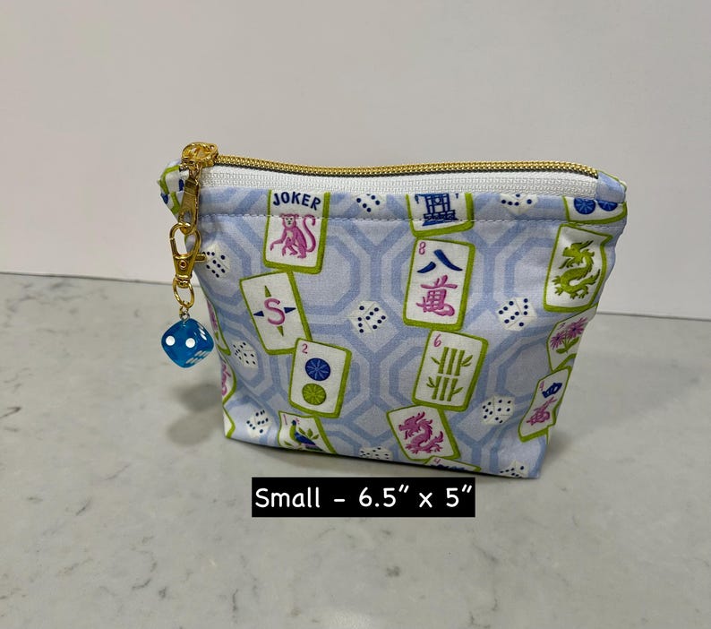 May include: Small, light blue zippered pouch with a gold zipper and a blue die charm. The pouch features a pattern of Mahjong tiles and dice. The pouch measures 6.5 inches by 5 inches.