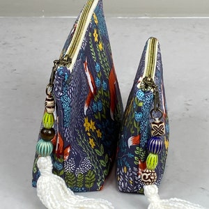 May include: Two triangular-shaped cosmetic bags with a navy blue fox and floral print. The bags have brass zippers, beaded zipper pulls, and white tassels. The bags are different sizes, suitable for travel or everyday use.