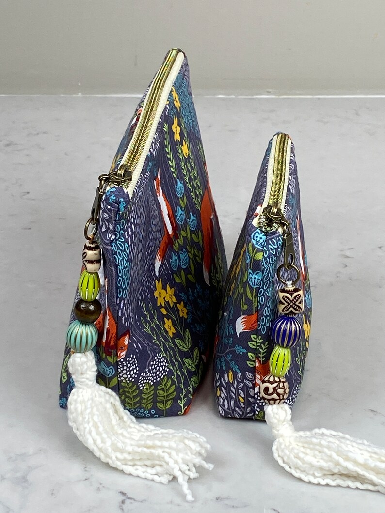 May include: Two triangular-shaped cosmetic bags with a navy blue fox and floral print. The bags have brass zippers, beaded zipper pulls, and white tassels. The bags are different sizes, suitable for travel or everyday use.