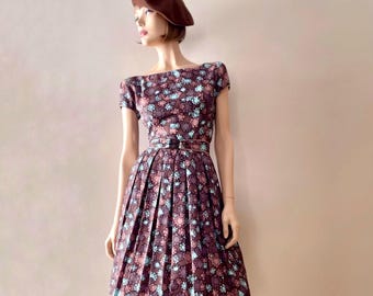 May include: A knee-length dress with a dark brown base featuring a repeating floral pattern in shades of light blue, pink, and white. The dress has short sleeves, a boat neckline, a pleated skirt, and a matching brown belt at the waist.
