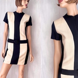 Puede incluir: Un minivestido vintage con diseño de bloques de color. El vestido presenta una combinación de colores negro y crema con un patrón de rayas verticales. Tiene mangas cortas, cuello alto y un cinturón negro que ciñe la cintura. El vestido se exhibe en un maniquí.