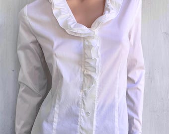 May include: A long-sleeved white button-down shirt featuring a ruffled collar that extends down the front placket. The shirt has fitted long sleeves with ruffled cuffs and a structured silhouette, crafted from a smooth, solid white fabric.