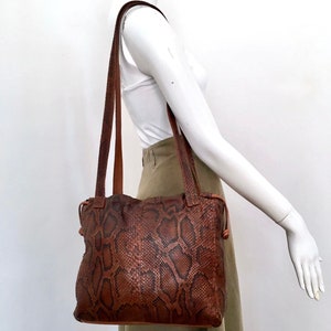 Może przedstawiać: Brązowa skórzana torba typu tote z wzorem skóry węża. Torba ma długi pasek i sznurek do zamykania.