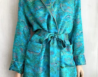 May include: A turquoise robe featuring a repeating landscape print with trees, pagodas, and bridges in shades of green, pink, and blue. The garment includes a shawl collar, long sleeves with cuffs, two front pockets, and a matching tie belt.