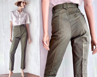 May include: High-waisted olive green trousers featuring a structured waistband with belt loops, a front zipper closure, and straight-leg tailoring with cuffed hems. The pants are shown on a mannequin paired with a short-sleeved white button-down shirt.