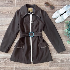 Peut inclure: Une veste marron foncé dotée d'une fermeture éclair blanche sur le devant, d'une ceinture assortie avec une boucle circulaire et de deux poches à rabat. Le vêtement présente des manches longues, un large col et des surpiqûres blanches contrastées.