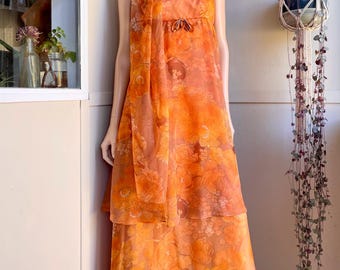 May include: A floor-length orange dress featuring a sheer overlay with a floral pattern, thin spaghetti straps, and a matching scarf draped around the neck. The bodice includes a small bow detail at the center of the empire waistline.