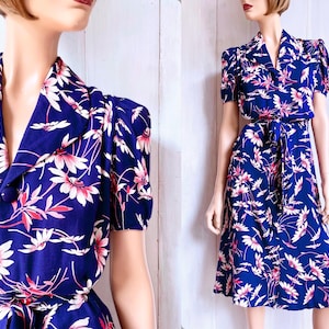 Könnte beinhalten: Vintage-Kleid mit marineblauem Hintergrund und floralem Muster aus weißen und rosa Blüten. Das Kleid hat kurze Ärmel, einen Kragen und einen passenden Bindegürtel. Das Kleid ist knielang.