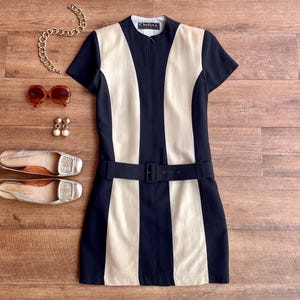 May include: A black and cream striped dress with a black belt, sunglasses, gold chain, earrings, and silver shoes. The dress has a Chelsea label. The items are arranged on a wooden surface.