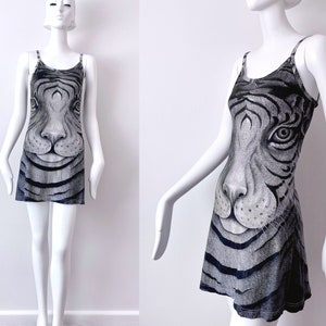 May include: A sleeveless mini dress featuring a detailed tiger face print in shades of gray and black. The dress has thin straps and a fitted bodice that flares out slightly at the hem. The tiger's face is centered on the front of the dress.